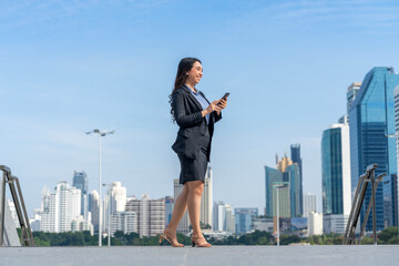 Confident Asian businesswoman using smartphone while walking in modern city. Professional female leader outdoors with skyscraper skyline.