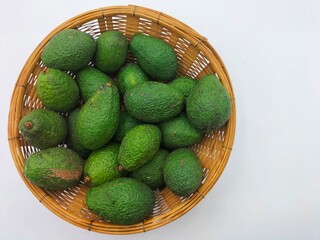 Several unripe, bright green Hass avocados are placed in a bamboo basket on a white background....