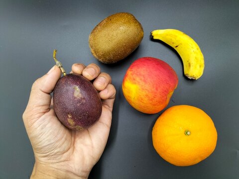 Five types of fruit are placed on a black background and are held in one's left hand. They include an apple, kiwi, banana, passion fruit, and orange.
