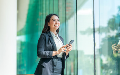 Fototapeta premium Asian businesswoman using smartphone near glass wall with reflection. Confident woman in formal outfit using phone in modern indoor space.