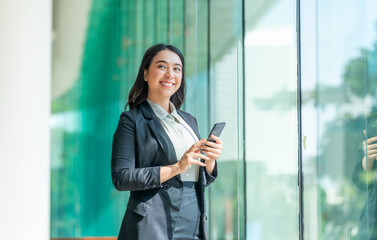 Asian businesswoman using smartphone near glass wall with reflection. Confident woman in formal...