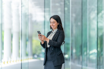 Fototapeta premium Smiling Asian businesswoman using smartphone in modern glass corridor. Confident woman in business attire standing indoors with mobile phone.