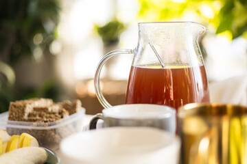 Close up of glass jug with fruit tea on cozy breakfast table with cups, granola bars and fresh fruit