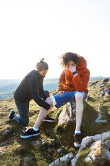 A person tends to another's injured knee outdoors. The injured person sits on a rock, while the...