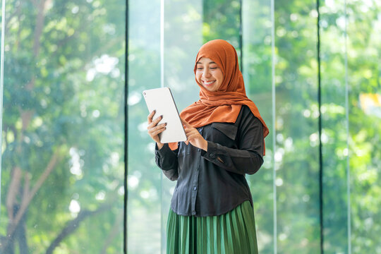 Smiling Asian muslimah using tablet indoors with natural outdoor view. Confident Muslim woman working on digital device near glass window. - Powered by Adobe