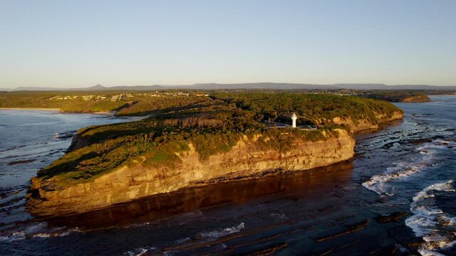 Warden Head Lighthouse stands proudly above rugged cliffs as waves crash far below. Framed by morning light, this coastal sentinel overlooks the vast, blue Tasman Sea near Ulladulla, NSW, Australia.