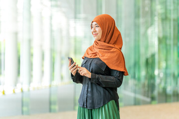 Smiling Asian muslimah using smartphone indoors with green outdoor view. Confident Muslim woman...