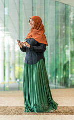 Smiling Asian muslimah using smartphone indoors with green outdoor view. Confident Muslim woman...