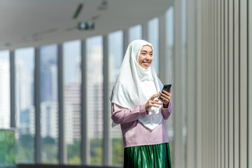 Smiling Asian muslimah using smartphone indoors with city skyline background. Confident Muslim...