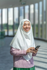 Smiling Asian muslimah using smartphone indoors with city skyline background. Confident Muslim woman using phone in a business office building.