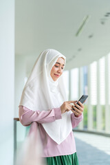 Smiling Asian muslimah using smartphone indoors with city skyline background. Confident Muslim woman using phone in a business office building.