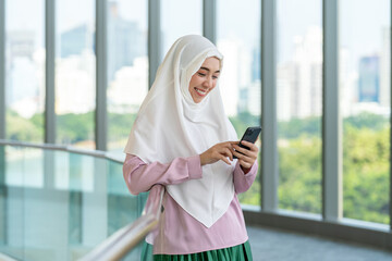 Smiling Asian muslimah using smartphone indoors with city skyline background. Confident Muslim...