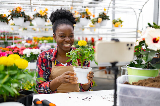 A smiling Black woman holds a potted plant, recording a video in a vibrant greenhouse filled with colorful flowers.