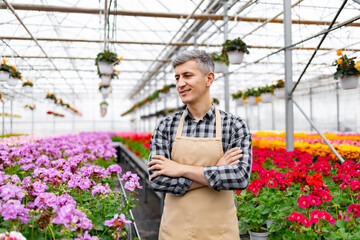 A smiling man with crossed arms stands in a sunlit greenhouse, surrounded by vibrant flowers and...