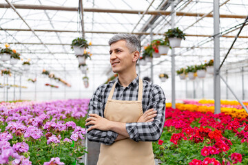 A smiling man in a greenhouse, surrounded by colorful flowers, wearing an apron and plaid shirt,...
