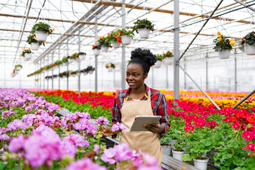An African-American woman, smiling, holding a tablet while inspecting colorful flowers in a...