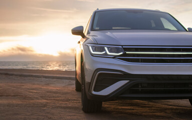 Silhouetted by a colorful sunset, a gray car stands near the ocean's edge. The warm light and open space capture a moment of solitude and exploration. 