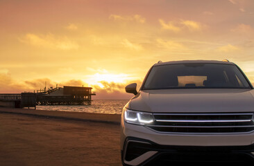 Silhouetted by a colorful sunset, a gray car stands near the ocean's edge. The warm light and open space capture a moment of solitude and exploration. 