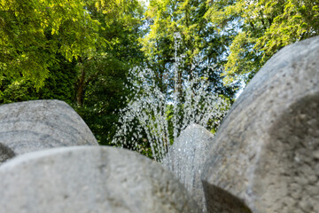 Water sprays from a stone fountain in a park surrounded by green trees. Shot from a low angle with...