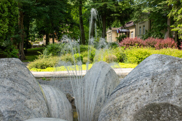 Fototapeta premium Outdoor stone fountain with water streams in a landscaped park surrounded by trees, bushes, and historic buildings in the background.