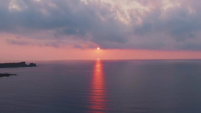 Setting sun casts a brilliant, fiery reflection path across a very calm sea, under a soft, cloudy sky. Distant land visible. Peaceful, serene seascape.