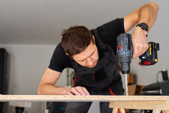Worker using power drill on wooden board supported by sawhorses in home garage workshop. Man working in workshop, drilling hole in chipboard for furniture assembly - Powered by Adobe