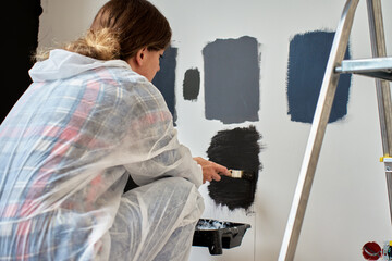 Woman in protective wear applies different dark paint swatches on white wall using brush. Female...