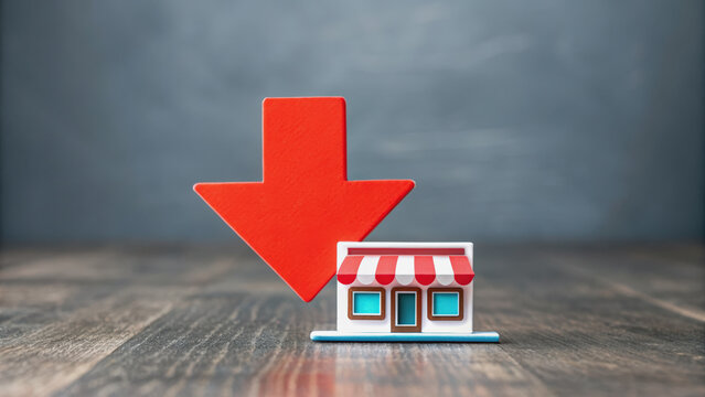 A miniature store icon with a large red downward arrow behind it on a wooden surface, symbolizing a decrease or decline in retail or small business.