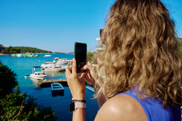 Rear view of woman in blue dress taking picture with smartphone near sea on sunny day. Female...