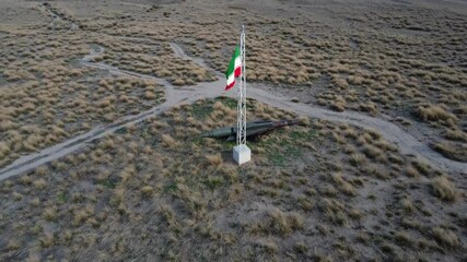 An Iranian flag stands tall next to a missile in a dry, grassy desert area, showcasing the blend of national symbolism and military presence in the remote location - Powered by Adobe