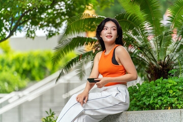 Young Asian woman using phone outdoors in urban green park. Beautiful young woman in casual...