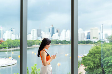 Young Asian woman using smartphone near large window with city view. Casual businesswoman using phone indoors.