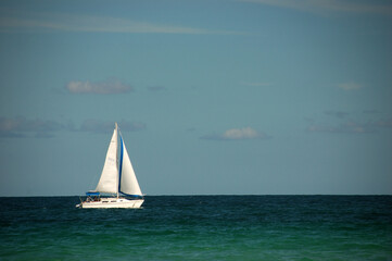 A white sailboat with blue accents sails peacefully on the turquoise ocean under a partly cloudy...