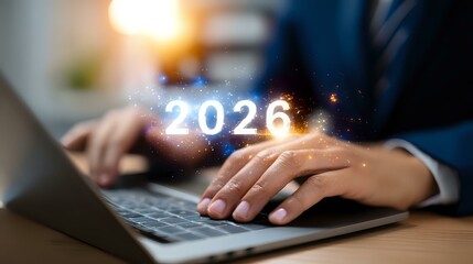 Glowing 2026 number floating above laptop keyboard with business person typing, magical light effect and sparkles in dark office environment.