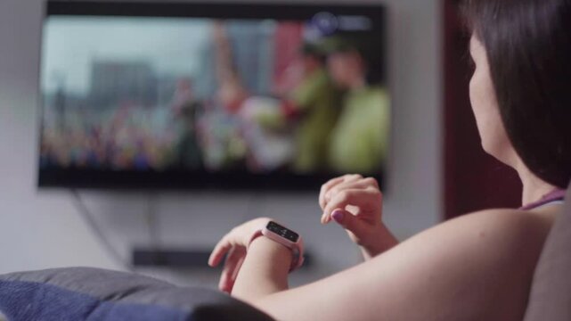 Woman relaxing on sofa, browsing smartwatch screen during tv program, representing contemporary connected digital living