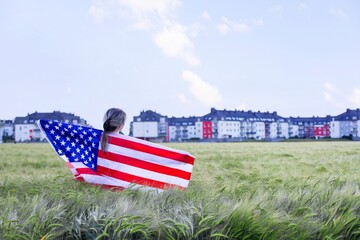 Woman with American flag in green barley field looking at city