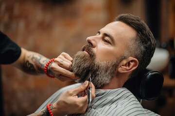 Professional barber trimming bearded man's facial hair with scissors in modern barbershop salon grooming service