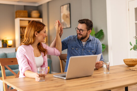 A happy man and woman high-five while working together at home. They smile, celebrating success or teamwork. A laptop and water glasses on the table suggest productivity in a cozy, modern setting.