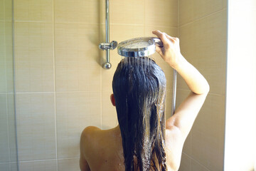 Woman washes her straight, medium-length, light brown hair under running water in shower, back...