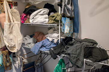 A cluttered home wardrobe with disorganized clothing, bags, and garments spilling out of shelves...