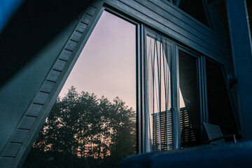 Close-up of a modern A-frame cabin window reflecting sunset and forest trees, with curtains partially drawn inside