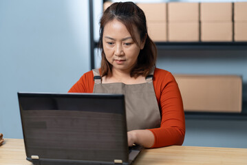 A dedicated Asian female entrepreneur in an apron works diligently on her laptop, managing her online shop and processing customer orders with inventory boxes visible in the background of her office.