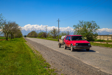 Red car pulling small trailer on rural road, sunny day in Kyrgyzstan