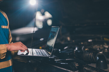 A mechanic uses a laptop to diagnose or repair a vehicle engine in a dimly lit garage or workshop.
