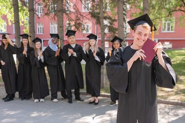 Young Caucasian man holding diploma while classmates applaud him. 