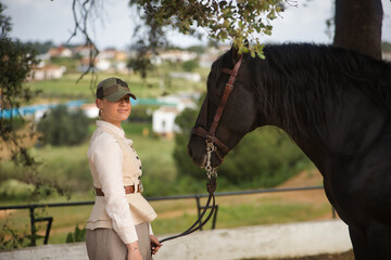 Young woman rider, posing next to her black Spanish thoroughbred horse, which she is holding by the...