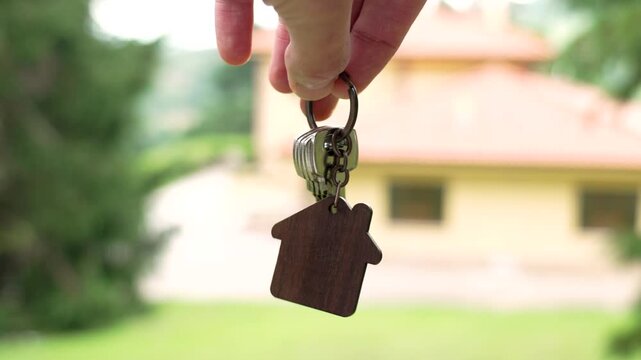 Home keys with wooden house shaped keychain against background of new bungalow in natural ecological area