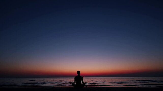 Silhouette of person meditating on beach at dusk with dark blue sky and ocean horizon, concept of self-care, mindfulness, mental health, yoga, and personal growth lifestyle content