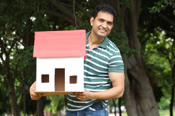 Indian happy young man holding 3D paper home shape model in park - Concept - Real Estate India