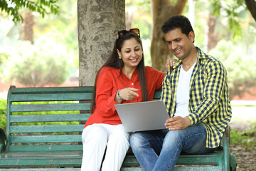 Indian happy couple working on laptop and doing online payment at park. Concept internet banking